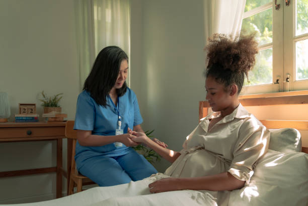 Asian home nurse female in blue gown measuring the pulsation heart rate of young African American pregnant woman who is laying down on bed at home. The family assistant nurse checking and following health of a pregnant woman monthly. enfermera asiática en el hogar con bata azul que mide la frecuencia cardíaca de la pulsación de una joven embarazada afroamericana que está acostada en la cama en casa. la enfermera asistente de familia revisa y sigue la salud de una mujer embarazada m