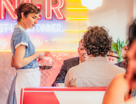 Profile view of a waitress in a traditional American diner as she offers coffee refills to customers.