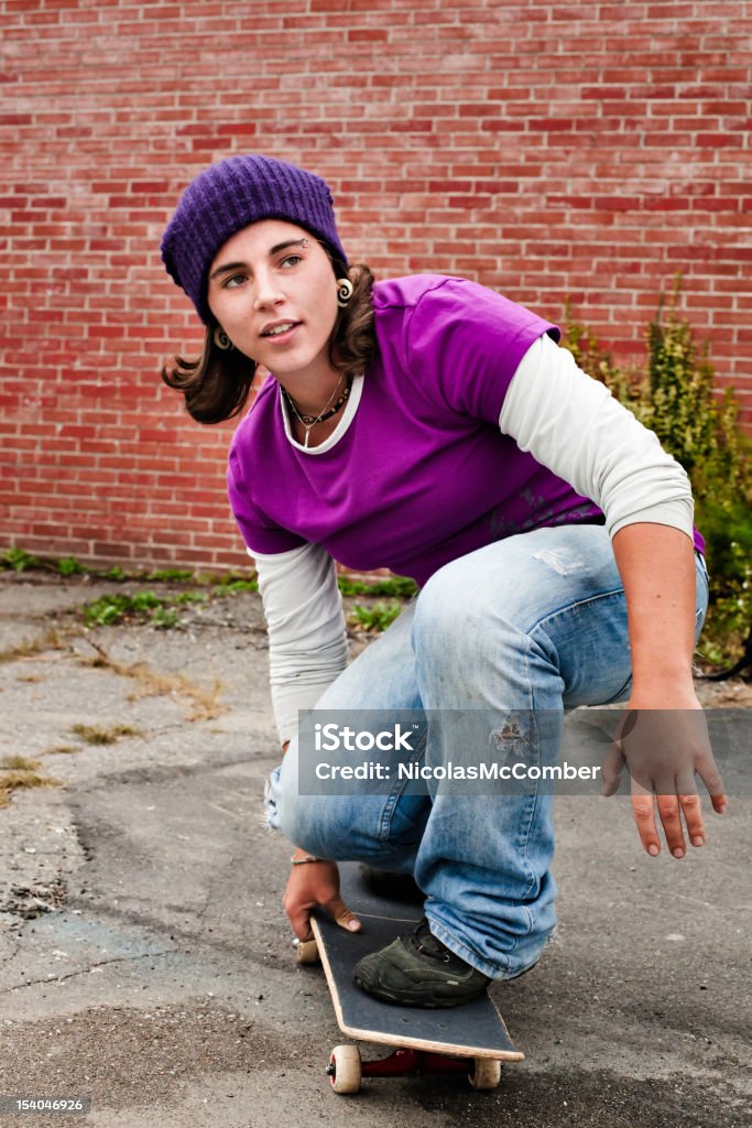 Skater girl on her skateboard Skater girl crouching on her skateboard in front of red brick wall. She is looking to the right with a relaxed smile. Purple Stock Photo