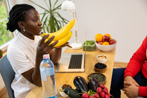 Female nutritionist of Black ethnicity and male client discussing a balanced nutrition plan 
On the desk, there are plenty of vegetables, fruits, dried fruits and nuts