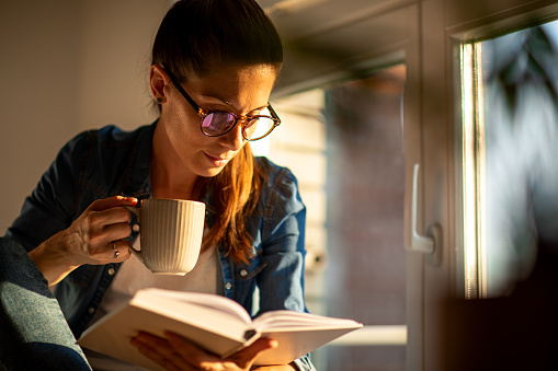 A woman sitting at her room window on at sunset and working on her laptop, listening music on her head phones and air pods, paying with credit card and texting on her smartphone.