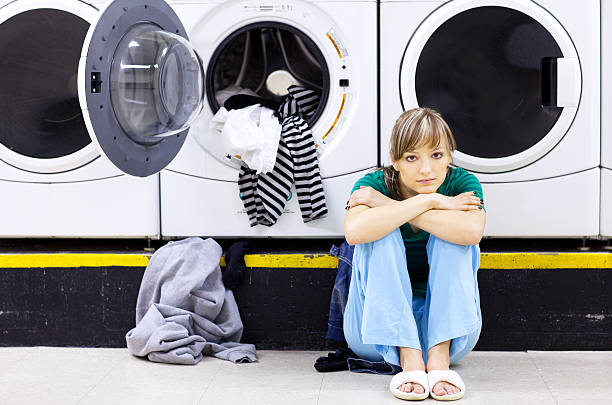 Tired woman doing laundry in laundromat, sitting on floor with clothes falling out.