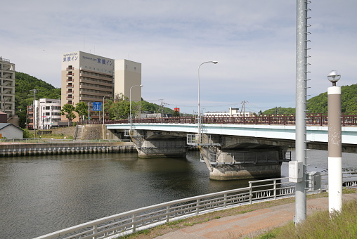 Abashiri City, Japan - June 1, 2023: The Shinbashi Bridge spans the Abashiri River. View looking southwest from the promenade across to the hotels in the Shinmachi area near the green hills and the scenic riverside. Spring morning with light clouds over northeastern Hokkaido Prefecture.