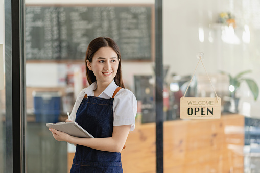 Asian woman standing by the open sign in front of the cafe door A beautiful smiling waitress with her arms crossed stands behind a doorplate in a cafe.