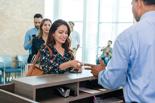 With a line of people waiting behind her, the mid adult woman smiles as she hands her debit card to the unrecognizable male bank teller.
