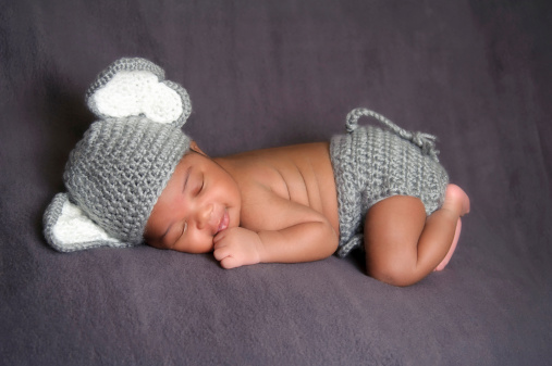 Thirteen day old smiling newborn baby boy wearing a gray crocheted elephant hat and diaper cover. He is sleeping on his stomach on gray fleece fabric.