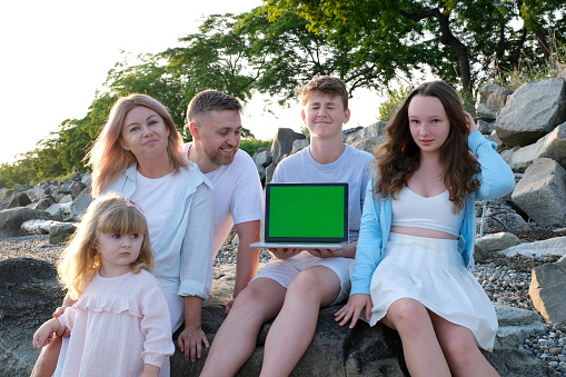 big family mom dad and three children boy girl teenagers baby look at the green screen and into the frame In the hands of a laptop with a place for advertising text sit on the rocks travel relaxation