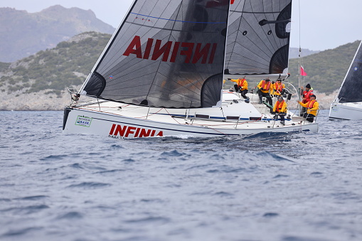 Bodrum,Mugla, Turkey. April 02,  2023: sailor team driving sail boat in motion, sailboat wheeling with water splashes, mountains and seascape on background. Sailboats sail in windy weather in the blue waters of the Aegean Sea, on the shores of the famous holiday destination Bodrum