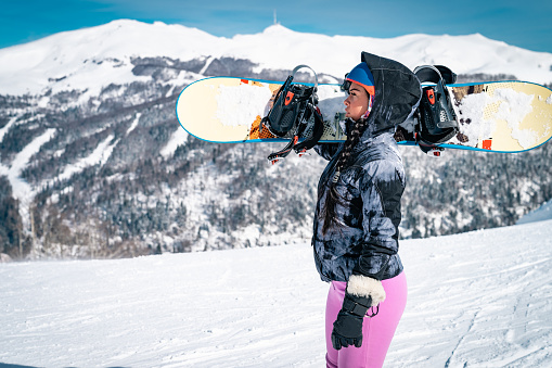 Young female snowboarder standing on top of mountain while holding a snowboard. Athletic young woman holding winter sport equipment outdoors.