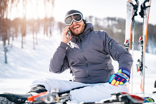 Smiling male skier sitting on snow taking a break while talking on mobile phone. Cheerful businessman having a call during his winter vacation in mountains.