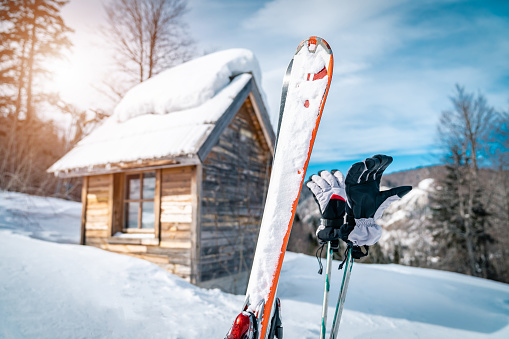 Close up of skis and ski gloves on ski poles on top of a mountain in front of log cabin. Idyllic winter scenery with wooden cottage covered with snow.
