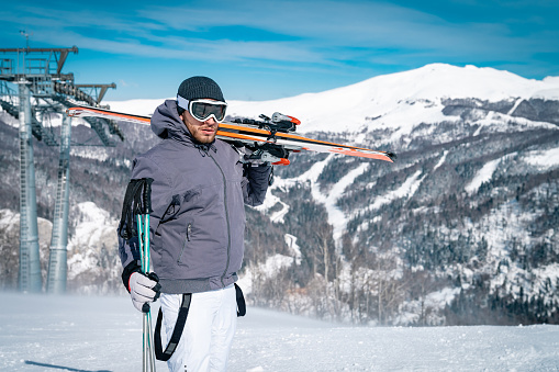 Young male skier standing on top of mountain while holding skies on his shoulder. Serious confident man standing outdoors in full winter sport equipment and looking at camera.