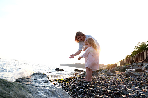 Mom and daughter stand on, beach with many stones at sunset and wet their hands from the sea ocean little cute girl and adult mom parents care love vacation family