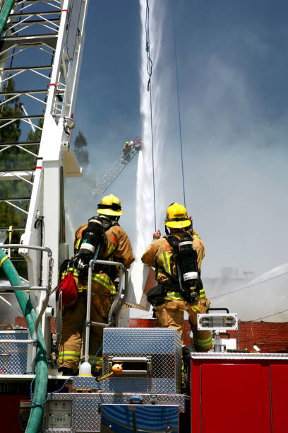 istock Firefighters standing on a hook and ladder fire engine watching as water canons blast a structure fire with streams of water. 1541055178