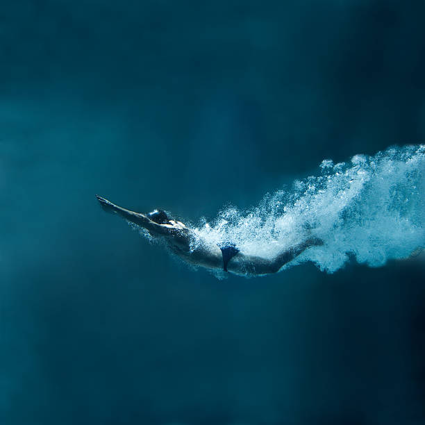 Swimmer underwater after the jump on blue background Male swimmer swimming dolphin. The image is made just after entering the water. His body is covered by air bubbles. Hands are joined in front of the body. He wears black cap and black swimwear. The swimmer has muscular built. He is leaving behind a lot of air bubbles. The background is dark blue. There are not visible parts of swimming pool. There is a lot of free space above and below him. jumping into water stock pictures, royalty-free photos & images