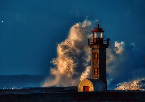 A large wave crashes against a pier in front of a picturesque lighthouse, illuminated by the setting sun