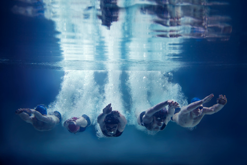 Five, young swimmers are jumping in direction of the camera. This is underwater shoot. There are 3 girls and 2 boys. They wear caps, swimming goggles and swimwears. The swimmers just entered the water with hands in front of themselves. They are looking down. They leave behind them a lot of air bubbles. In the top of image you can see the surface of water and reflection of the scene. The background is clear, dark blue. There are no swimming pool elements. This is a horizontal image.