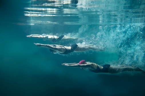 On the image there are 3 female swimmers. They are wearing black one-piece swimwears, cups and swimming goggles. The position of swimmers is straight, they have hands joined in front. Girls are looking down. There is a lot of air bubbles behind them. In the top of image there is water surface. Everything is on blue background. This is a horizontal photography.