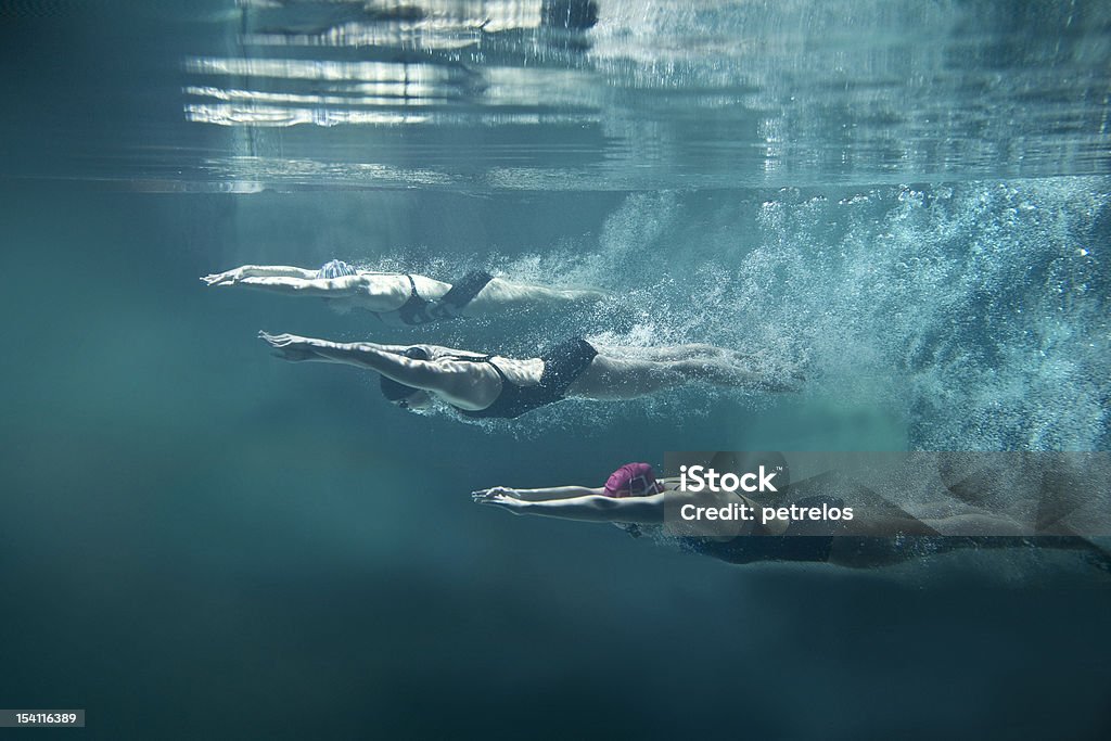 Three swimmers underwater divining after start On the image there are 3 female swimmers. They are wearing black one-piece swimwears, cups and swimming goggles. The position of swimmers is straight, they have hands joined in front. Girls are looking down. There is a lot of air bubbles behind them. In the top of image there is water surface. Everything is on blue background. This is a horizontal photography. Swimming Stock Photo