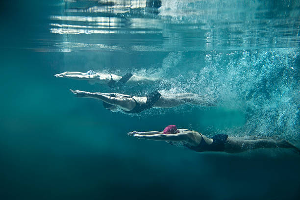 Three swimmers underwater divining after start On the image there are 3 female swimmers. They are wearing black one-piece swimwears, cups and swimming goggles. The position of swimmers is straight, they have hands joined in front. Girls are looking down. There is a lot of air bubbles behind them. In the top of image there is water surface. Everything is on blue background. This is a horizontal photography. team sport stock pictures, royalty-free photos & images