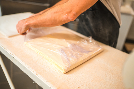 Close up on hands of Caucasian baker in 30s using a dough sheeter machine to produce laminated dough for croissants at the small artisanal bakery. He is wrapping laminated dough into a plastic foil.