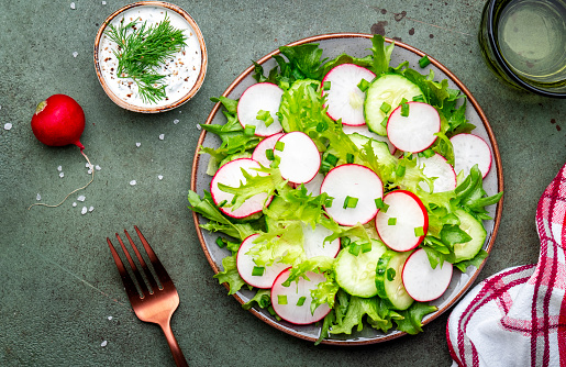 Healthy crispy salad with radishes, cucumbers, lettuce and chives with yogurt dressing, green stone table background, top view