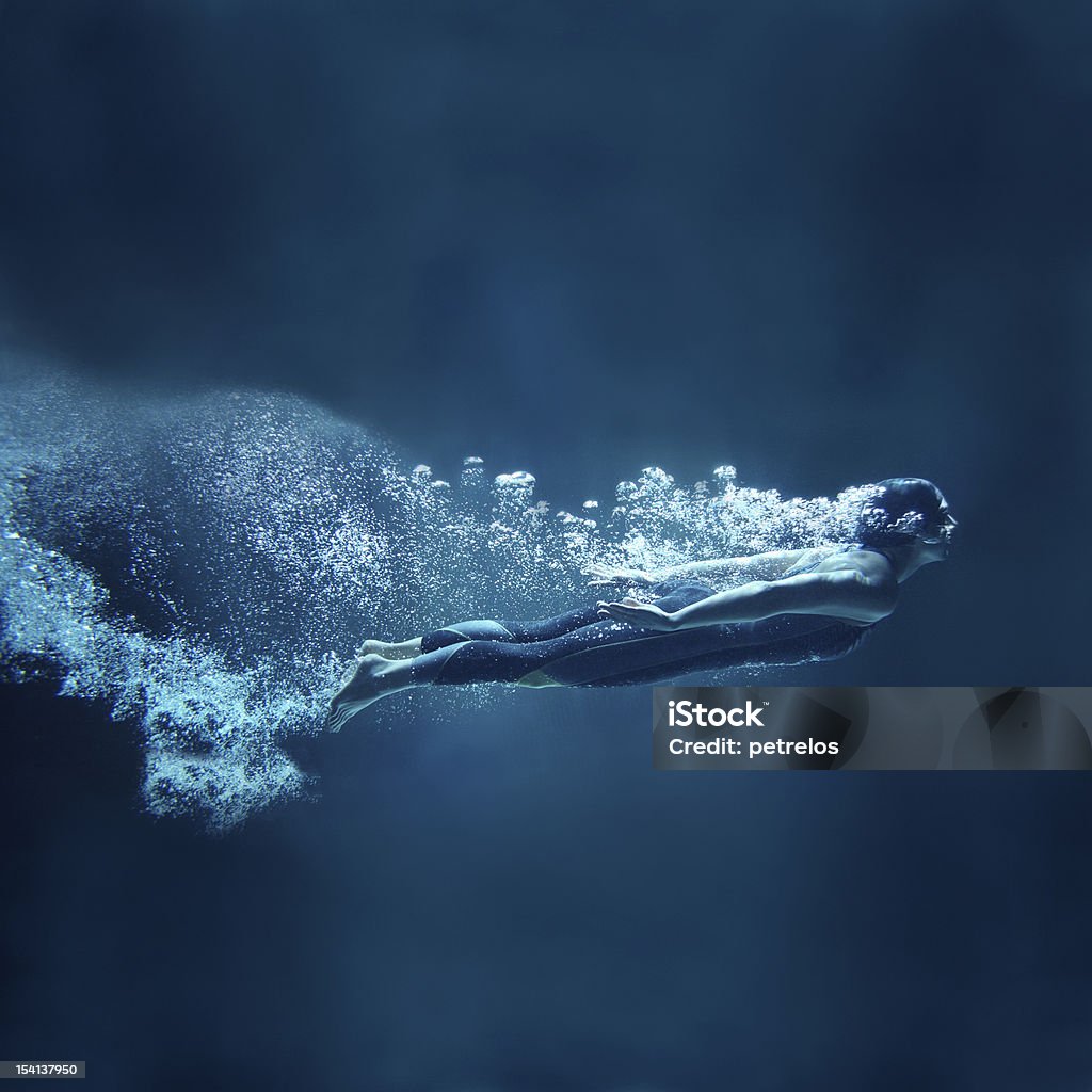 Female swimmer underwater flowing on blue background Athlete is dressed in a professional black swimwear. She is swimming horizontally like she was flying. She has two hands together along the body. She is looking ahead.  Behind her body you can see a lot of air bubbles. The background is dark blue- like open water. This is a square format photography. Women Stock Photo