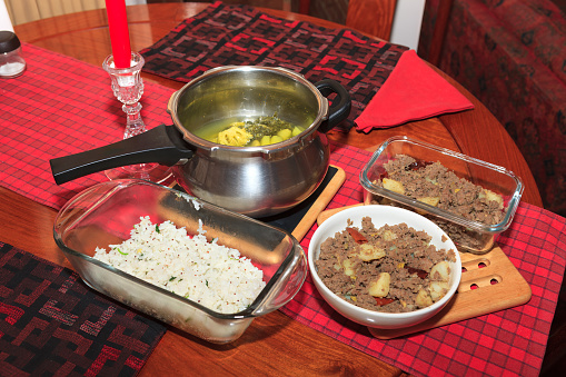 An Indian dinner is under preparation in a small Colombian apartment home. The finished dishes of Yogurt Rice and Minced Beef with Potato have been placed on a rosewood dinner table. The soup is yet to be blended and some other dishes are to be completed.