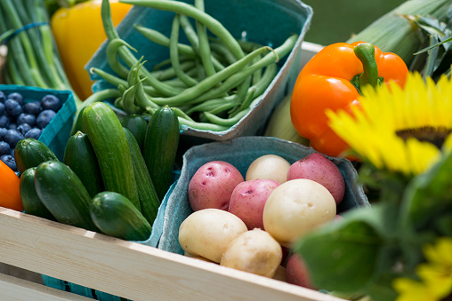 Closeup macro photo of a large arrangement of fresh, organic vegetables that are being sold at a farmers market booth. There is raw potatoes, zucchini, green beans, pepper, blueberries, asparagus, and sunflowers for purchase in a wooden crate.