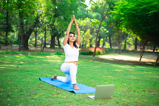 Young indian woman doing fitness stretching yoga exercise in the park while using laptop for online class or virtual tutorials. healthy lifestyle.