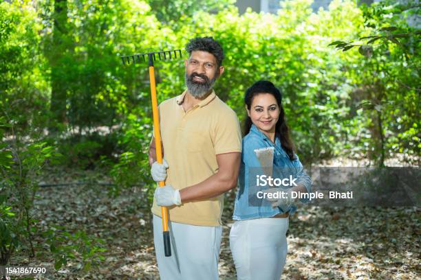 Portrait Of Happy Indian Couple Volunteer Holding Cleaning Equipments To Clean Fallen Leaves In The Forest Or Nature Spring Trash Cleaning In Park Looking At Camera Stock Photo - Download Image Now