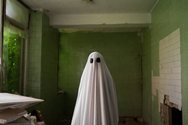 Ghost in a white sheet stands in an abandoned building against a peeling wall. Halloween concept. Copy space
