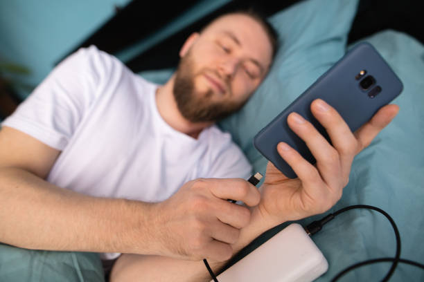 hombre millennial serio que usa el teléfono móvil en la cama lo conecta a un cargador mientras está acostado de lado - bedroom authority bed contemporary fotografías e imágenes de stock