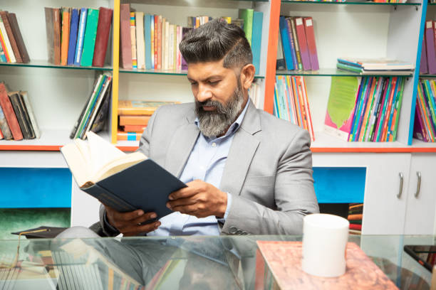 istock Smart mature indian man wearing suit reading a book while sitting at desk in library with bookshelf behind him, research, knowledge and learning concept 1541880487