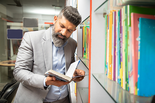 Smart mature indian man wearing suit reading a book at library standing by bookshelf, research, knowledge and learning concept