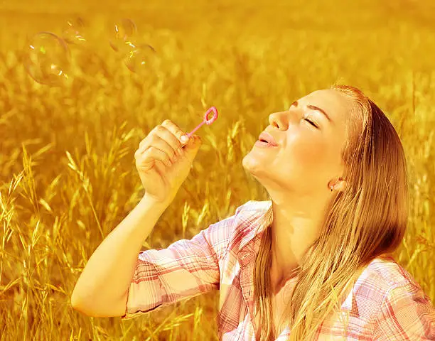Image of cute blond girl blowing soap bubbles on wheat field, happy teenager having fun on golden crop meadow, closeup portrait of pretty young woman playing game on farm land, autumn season