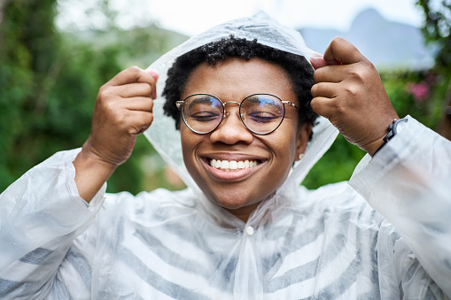 Close-up portrait of a happy young african woman putting on raincoat cap standing outside in rain