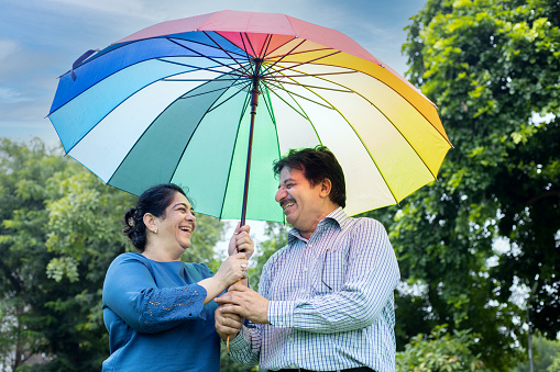 Happy senior indian couple standing under big multicolor or colorful umbrella at summer park. husband and wife. Life and health insurance Safety concept. low angle shot.
