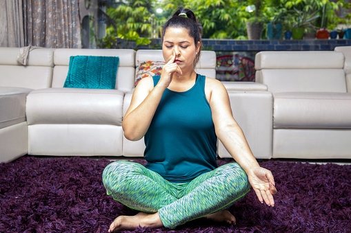 Young indian woman in sportswear sitting in Sukhasana pose, Alternate Nostril Breathin exercise at at home. Asian Urban Female doing yoga pranayama. Full length shot.