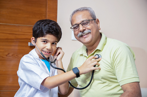 Playful indian boy kid wearing doctor uniform dress standing doing check heartbeat of with his grandfather with stethoscope at home. Closeup