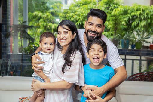 Cheerful Young Indian Family Of Four Having Fun Together At Home. Portrait Of Smiling Parents Playing With Their Children Looking At Camera.