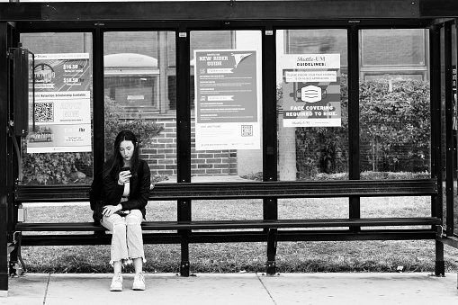 College Park, MD, USA-March 05, 2022: Women sitting on a bus stop bench looking at phone on the University of Maryland campus