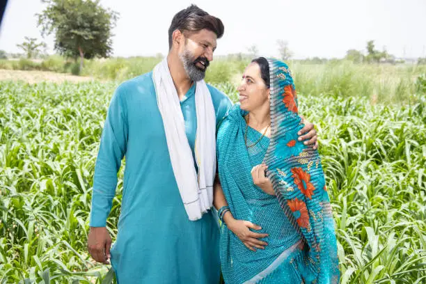 Joyous Indian couple farmers looking at each other with love in agriculture field, smiling rural beard man and woman standing together in farm land, village husband and wife enjoy relax in cropland.