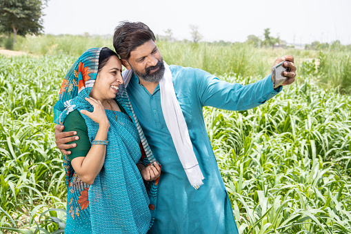 Joyous Indian couple farmer taking selfie on smart phone stand in agriculture field, rural man woman holding mobile phone on farmland, male female enjoying with cellphone outside, Digital India concept.