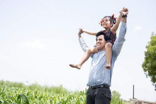 Happy Indian cute little adorable daughter sitting on father shoulder having fun in field, spend leisure time together in farm, girl child enjoy childhood with man outdoor, parent child relationship.