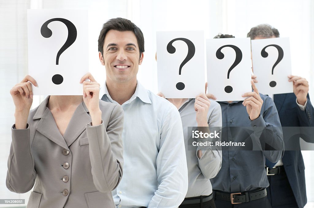 Row of people holding question marks over their faces Happy smiling business man standing out of the crowd with other people hiding their face behind a question mark sign. A Helping Hand Stock Photo
