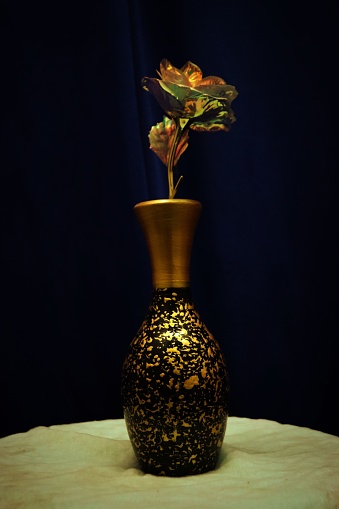 A metallic vase with a flower prominently displayed on a table in a dimly lit room.