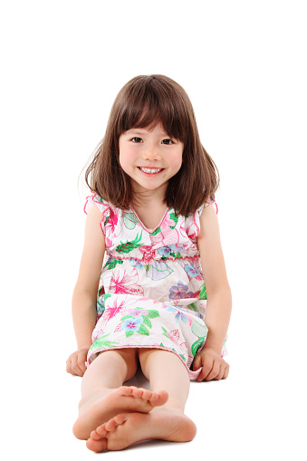 Young girl in a sun dress smiles at the camera as she sits on the floor. Vertical shot. Isolated on white.