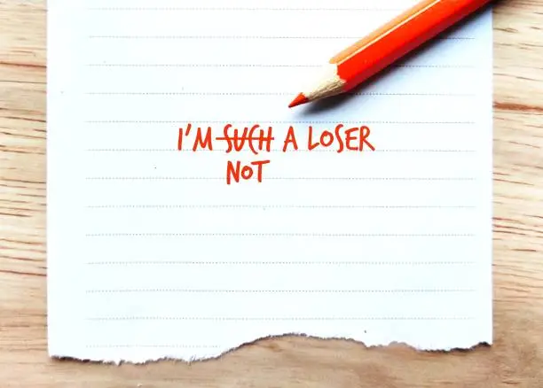 Orange pencil on note paper with text written I AM SUCH A LOSER, and change to I AM NOT A LOSER, concept of boost up low self esteem - turning negative self-talk into something more positive.
