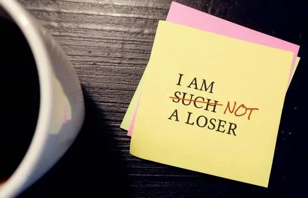 Yellow note paper with text written I AM SUCH A LOSER crossed off to I AM NOT A LOSER, to boost up low self esteem - turning negative self-talk into something more positive.